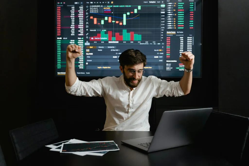 a person sits in front of a laptop with a large screen behind them showing active trading of L&T Finance share price. The person appears pleased with the ongoing activity.