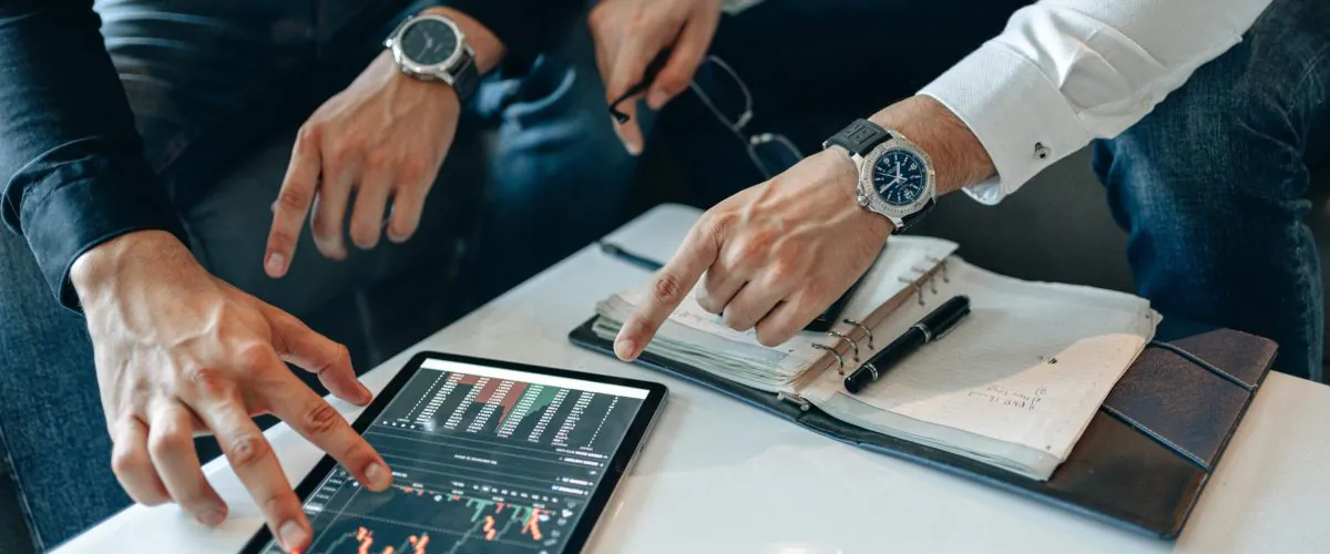 A person is pointing at a tablet showing investment activities, resembling participation in stock market classes at a training center focused on the stock market.